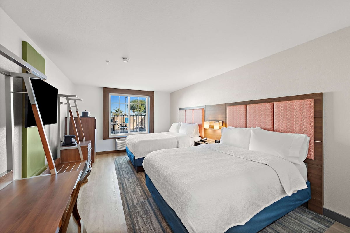 A hotel room with two beds, a desk, a chair, a TV, and a window with a view of palm trees.