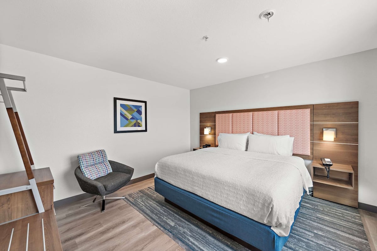 A modern hotel room with a large bed, a chair, a desk, and a painting on the wall.