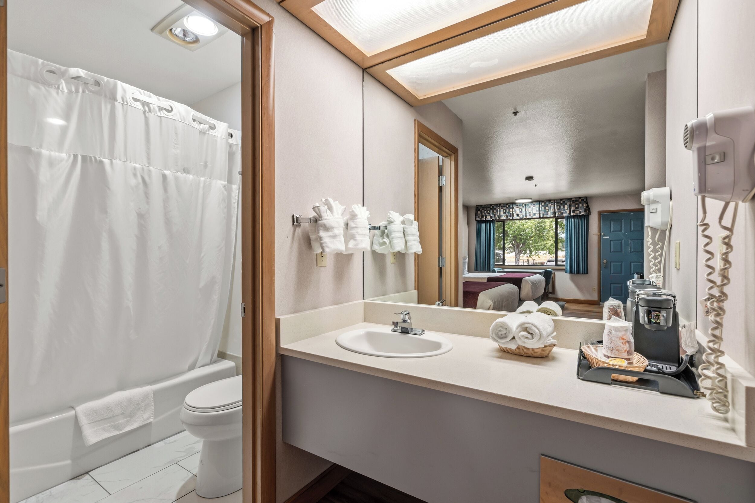 Baño con ducha, inodoro, lavamanos y espejo que refleja una cama y ventana.