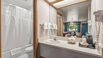 Baño con ducha, inodoro, lavamanos y espejo que refleja una cama y ventana.