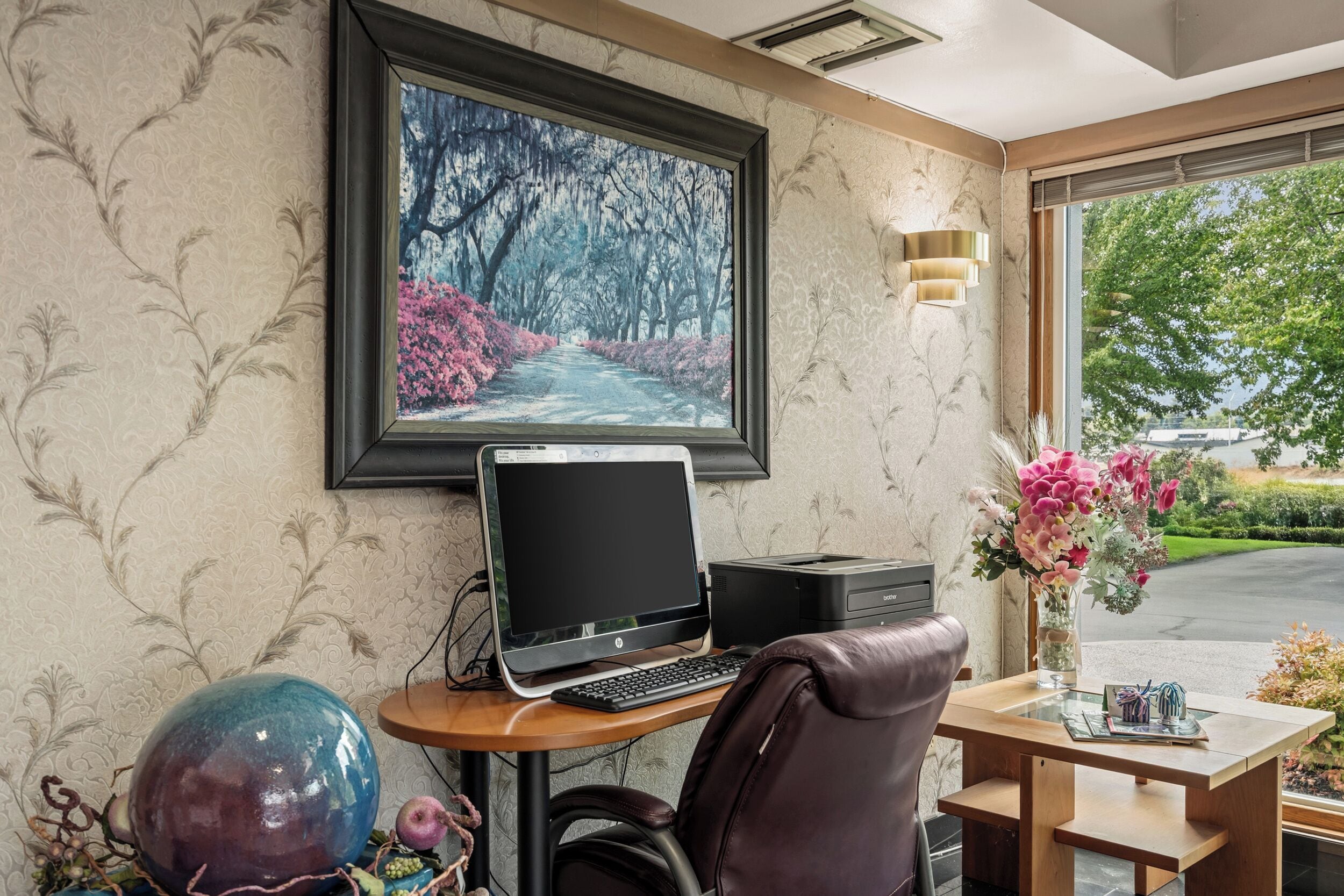 A hotel room with a desk, a laptop, a printer, a framed picture of a tree-lined path, and a vase with flowers.