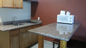 A kitchen with wooden cabinets, a granite countertop, a stainless steel sink, and a white microwave. - Scottish Inn & Suites (Houston)