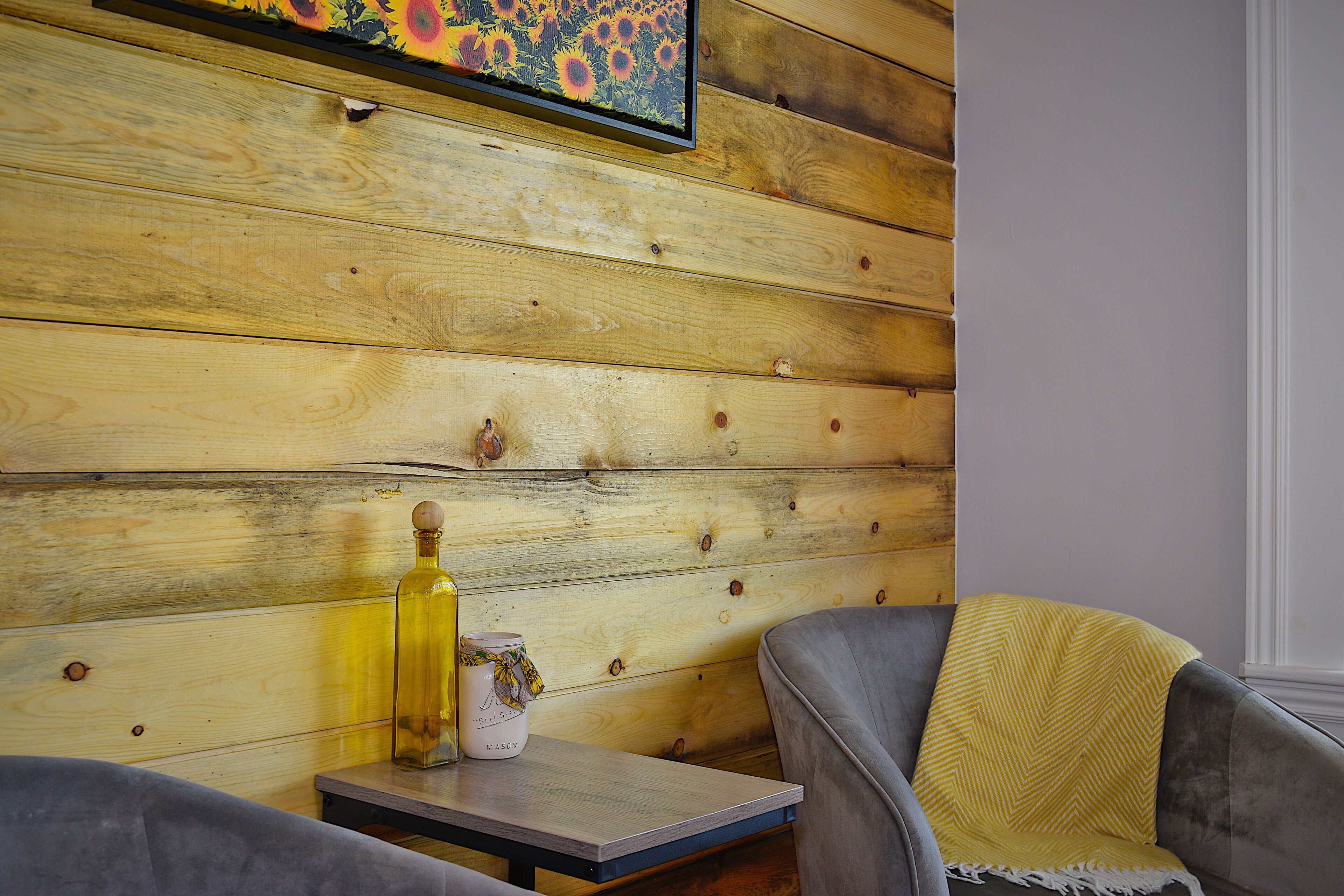 Ein Zimmer mit Holzwandverkleidung, einem kleinen Tisch mit einer Flasche und einem Dekorationsgegenstand sowie einem grauen Sessel mit gelbem Überwurf.