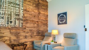 Una habitación con dos sillones grises, una pequeña mesa blanca con una lámpara, una pared con una obra de arte alusiva al café y una puerta.