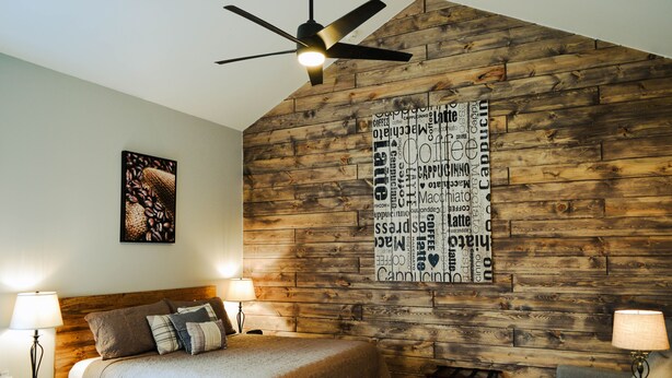 Un dormitorio con pared de madera, ventilador de techo, cama con manta, dos mesitas de noche con lámparas y una pequeña mesa con una planta.