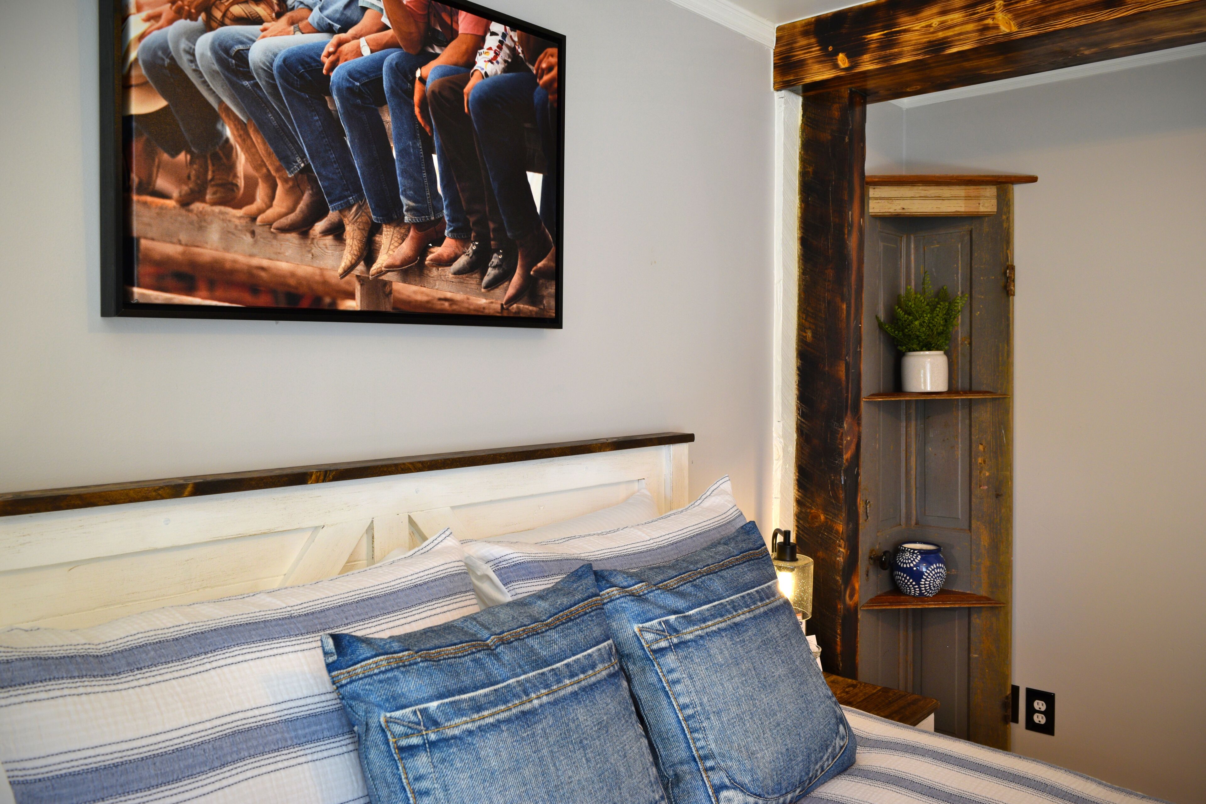 A bedroom with a bed, a framed picture on the wall, a wooden door, and a shelf with a plant.