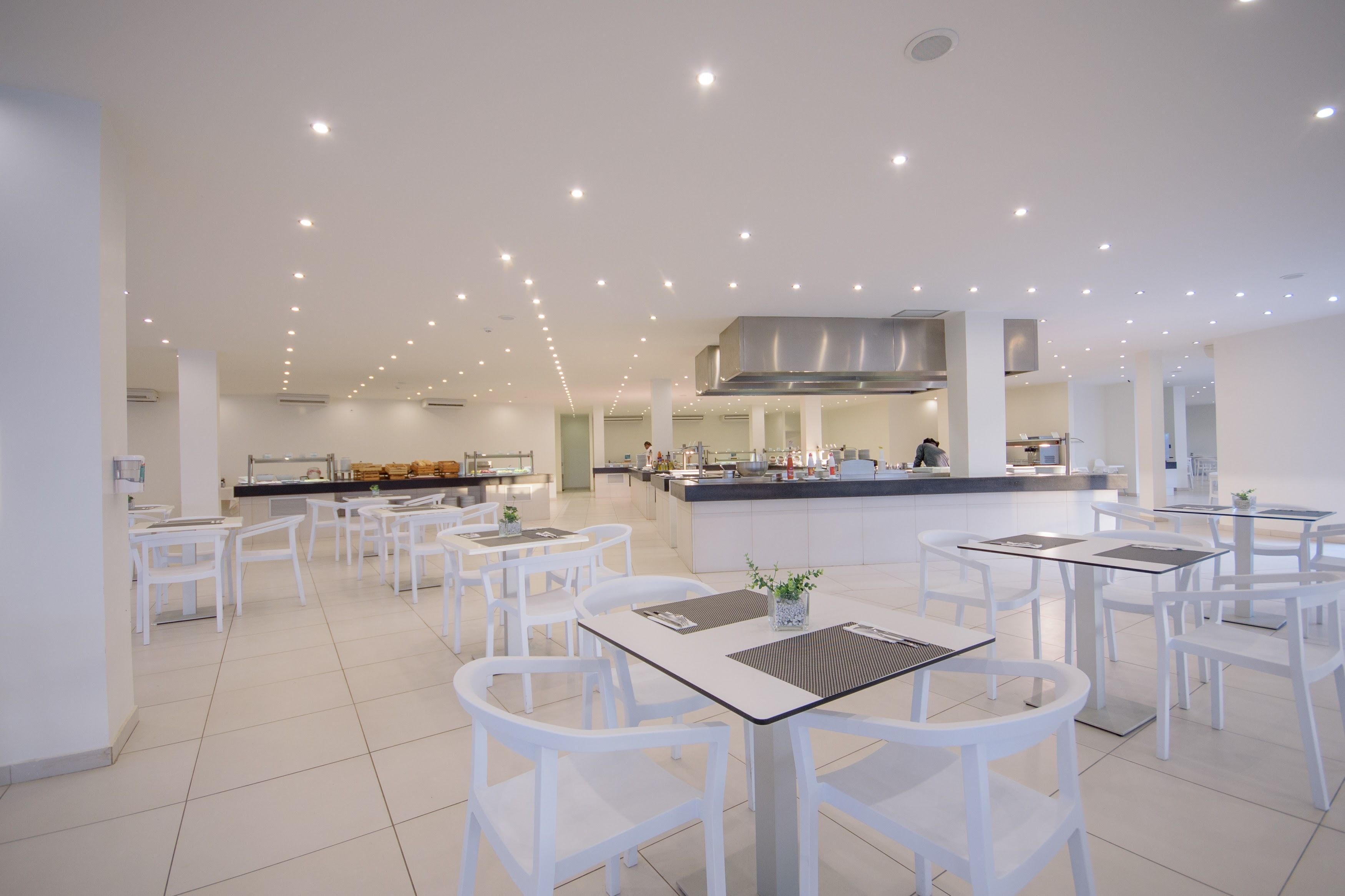Una sala da pranzo moderna e luminosa, con tavoli e sedie bianche, una zona cucina centrale e un design pulito e minimalista.