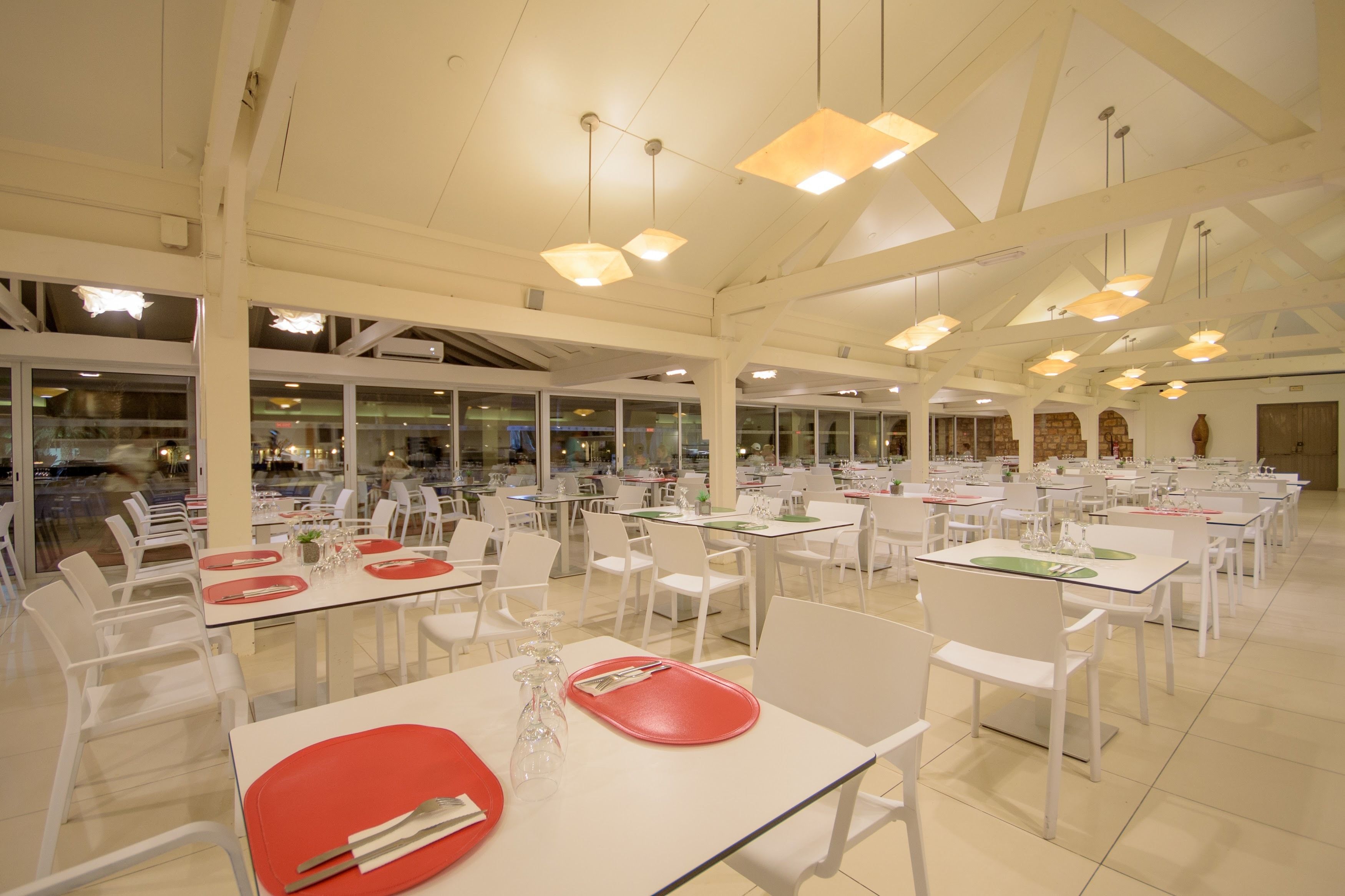 Una cafetería espaciosa con mesas y sillas blancas, ventanas amplias y lámparas colgantes.