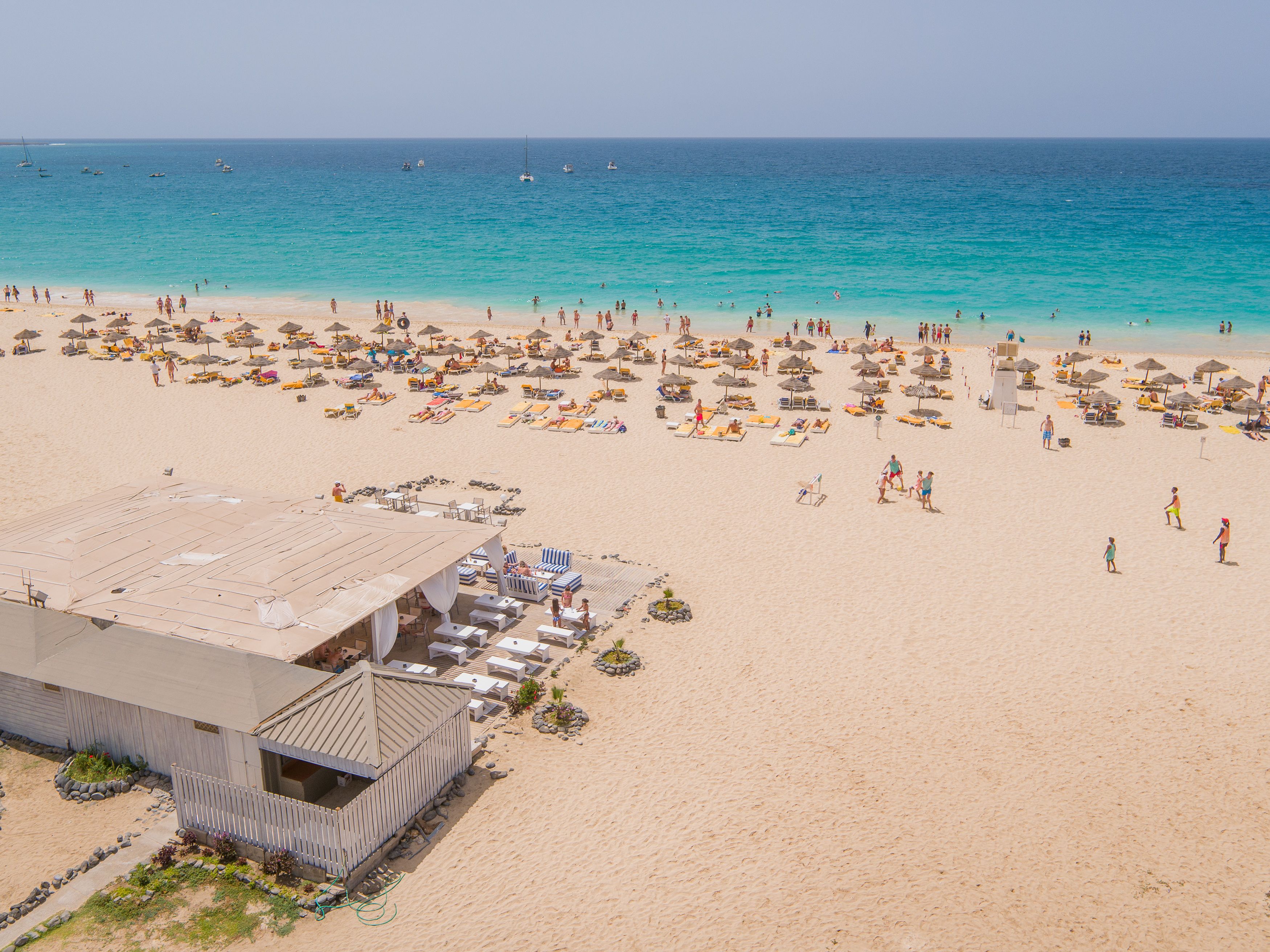 A beach with numerous sun loungers and umbrellas, a building with a flat roof, and a clear view of the ocean.