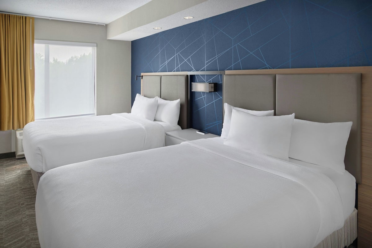 Habitación de hotel con dos camas, un ventanal con cortinas, un diseño geométrico azul en la pared y un cabecero moderno.