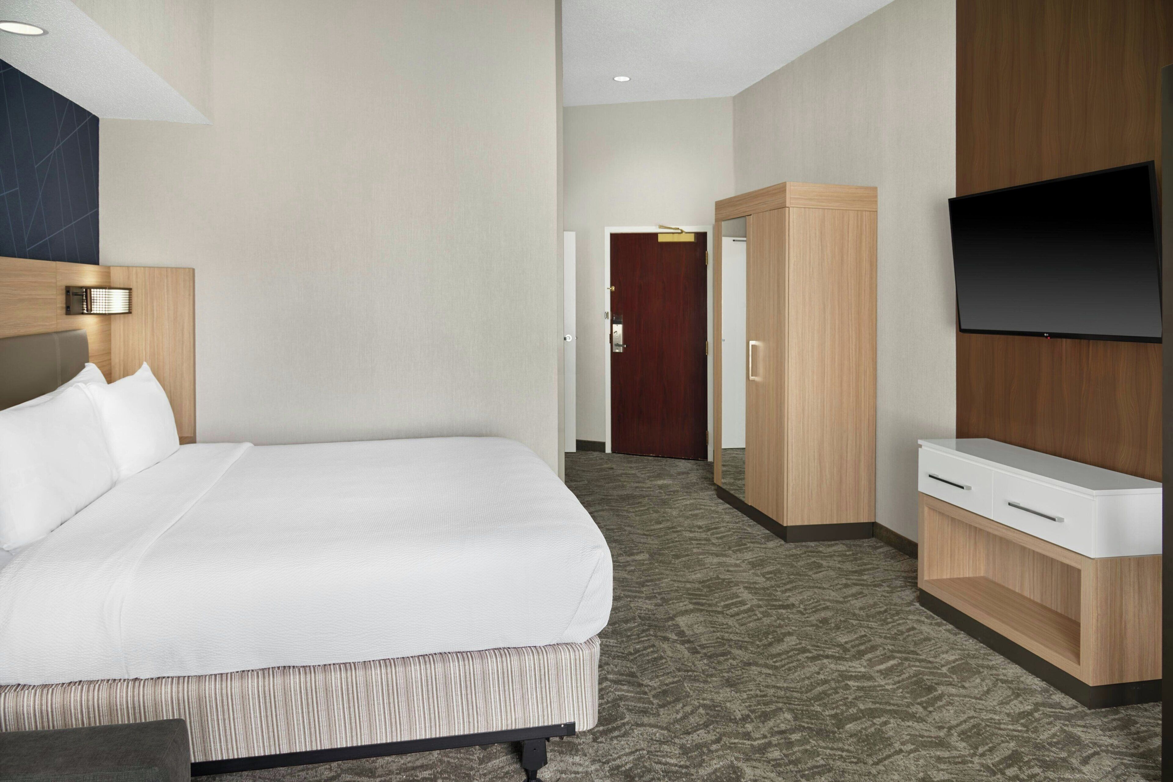 Une chambre d’hôtel avec un grand lit, un téléviseur fixé au mur, une armoire en bois et un sol recouvert de moquette.