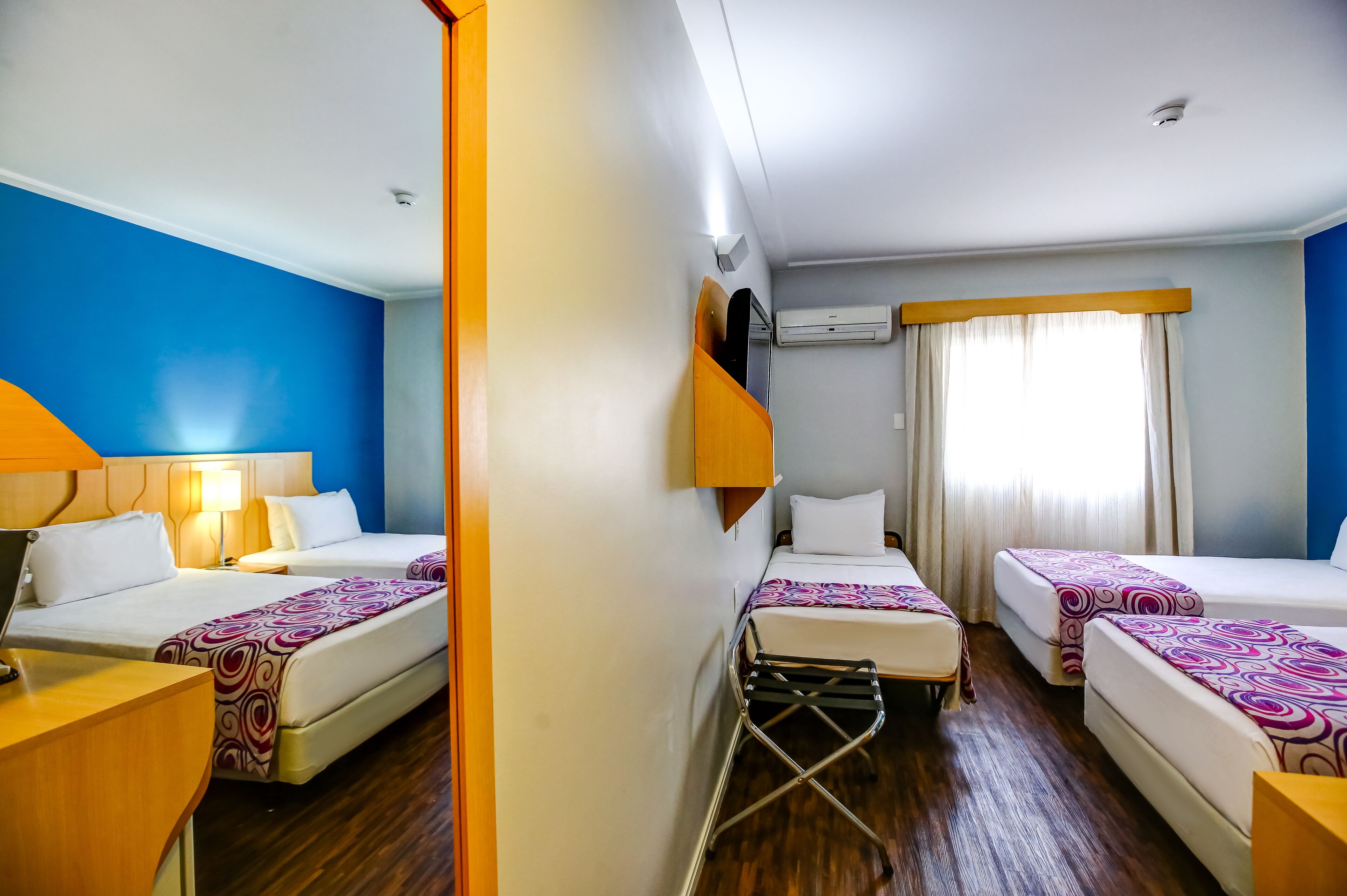 A hotel room with two single beds, a desk, a chair, an air conditioning unit, and a window with curtains.