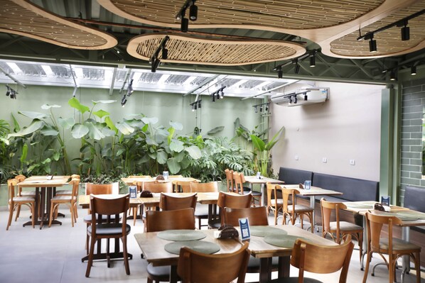 Um restaurante com mesas e cadeiras de madeira, plantas verdes exuberantes e teto alto com vigas de madeira circulares.