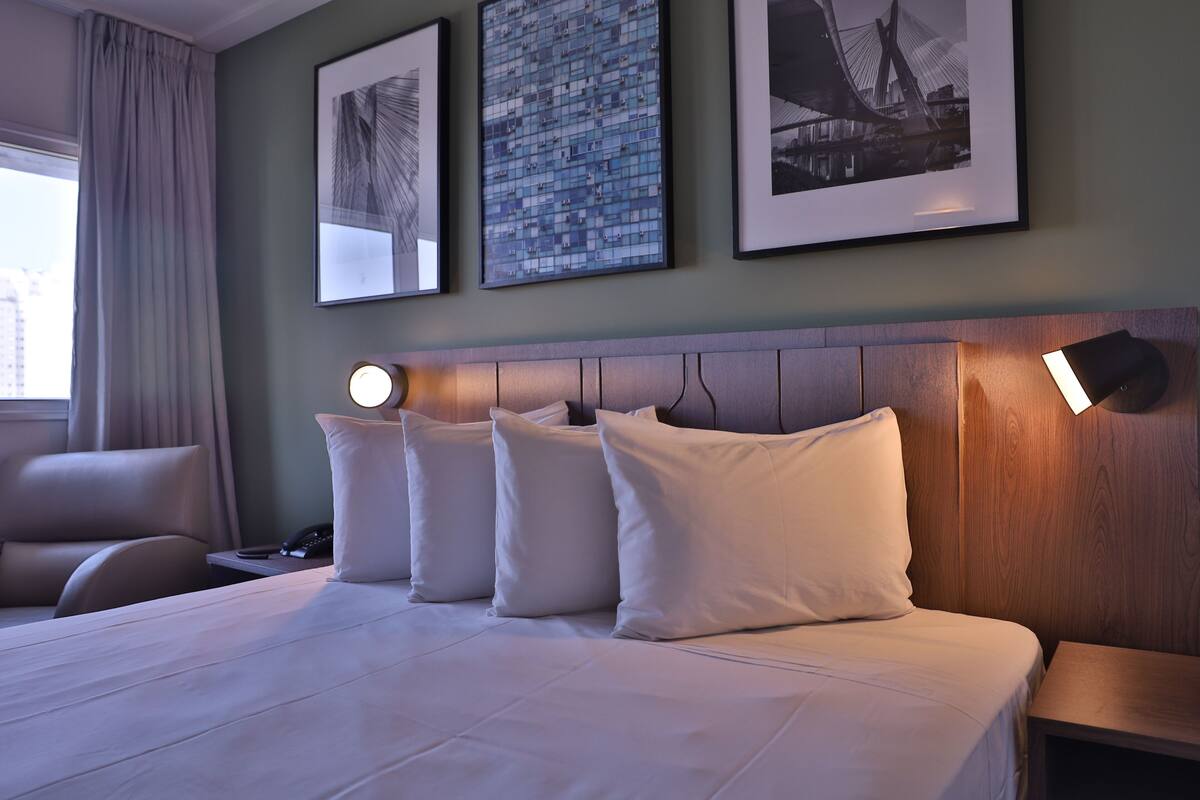 Une chambre d’hôtel avec un grand lit, une tête de lit en bois et trois lampes de chevet. Trois tableaux encadrés sont accrochés au mur, au-dessus du lit.