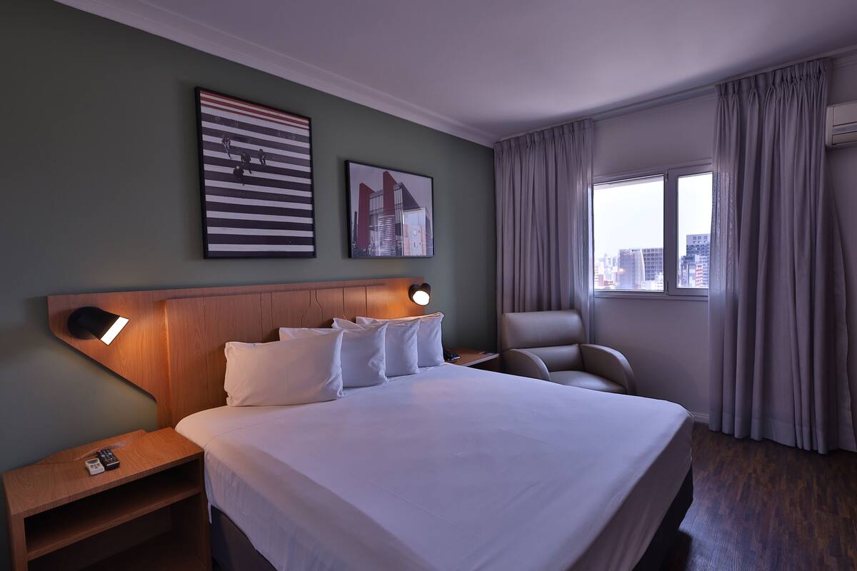 Une chambre d’hôtel avec un grand lit, une table de chevet avec une lampe, une chaise et une fenêtre avec des rideaux.