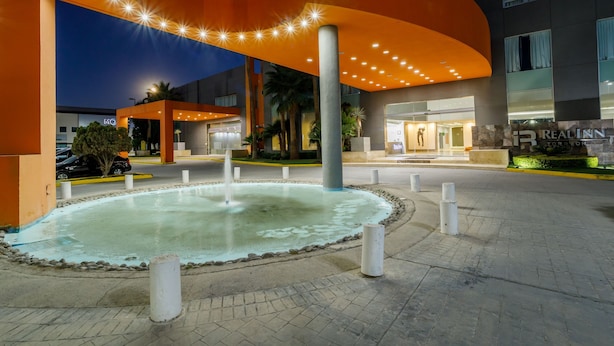 A modern hotel entrance with a central fountain, a curved orange canopy, and a well-lit facade.