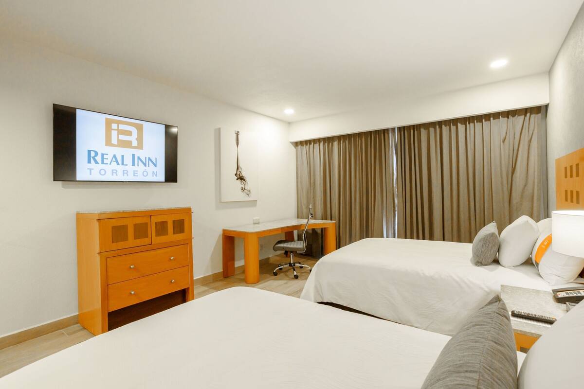 A hotel room with two beds, a desk, a chair, and a television displaying the Real Inn Torreon logo.