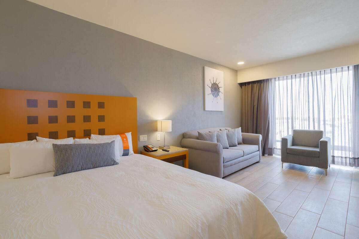 A hotel room with a large bed, a sofa, a chair, and a small table with a lamp.