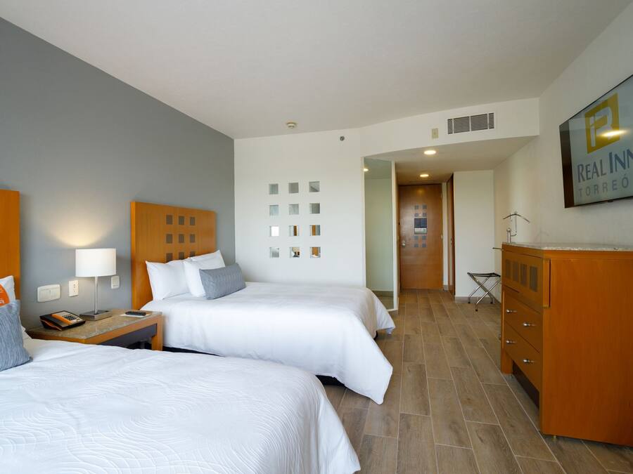 A hotel room with two beds, a wooden dresser, a desk with a lamp, and a wall-mounted TV.