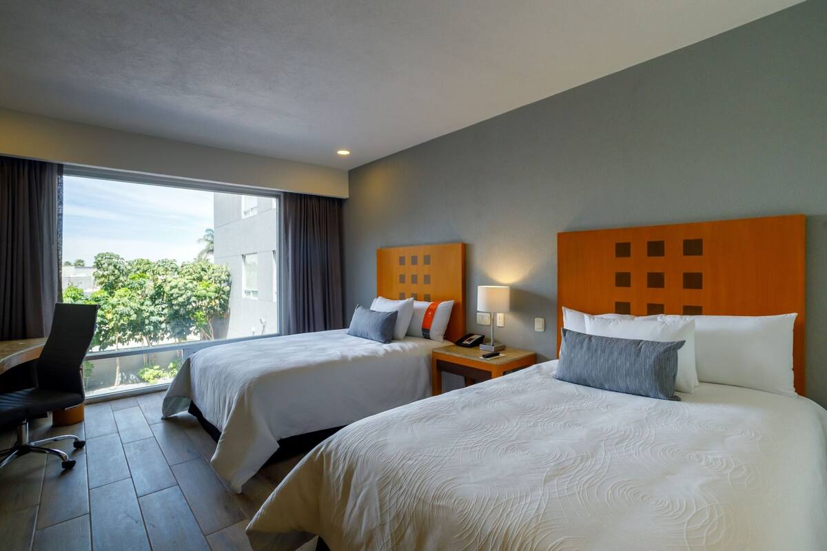 A hotel room with two beds, a desk, and a window with a view of greenery.