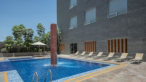 A swimming pool with a water feature, surrounded by lounge chairs and a modern building with large windows.