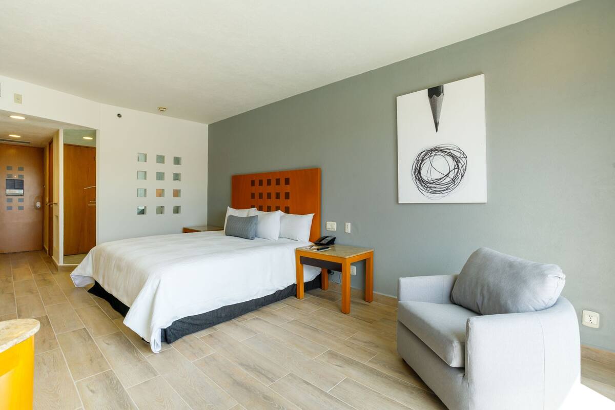 A hotel room with a bed, a wooden nightstand, a grey sofa, and a wall with a circular abstract artwork.