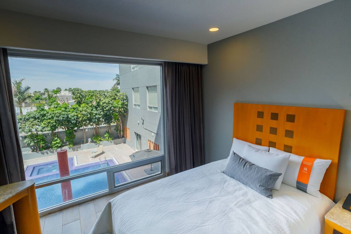 A modern hotel room with a large bed, a view of a swimming pool, and a cityscape visible through the window.