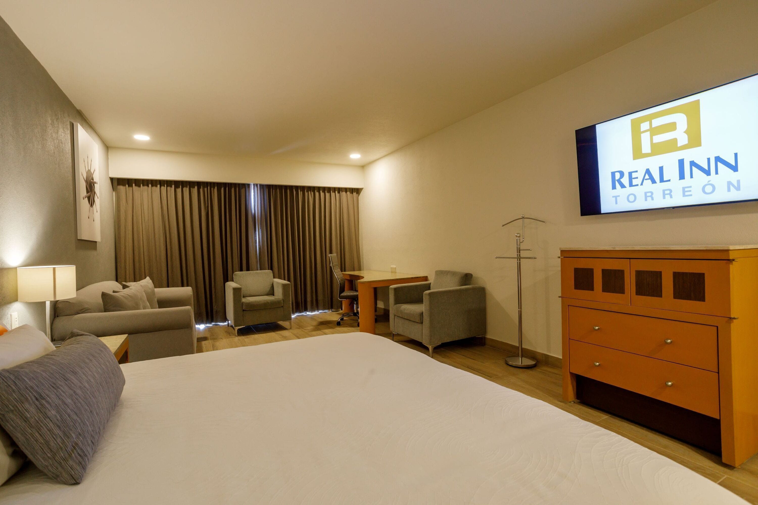 Une chambre d’hôtel avec un grand lit, deux fauteuils, un bureau, une commode et un téléviseur affichant le logo de l’hôtel Real Inn Torreón.