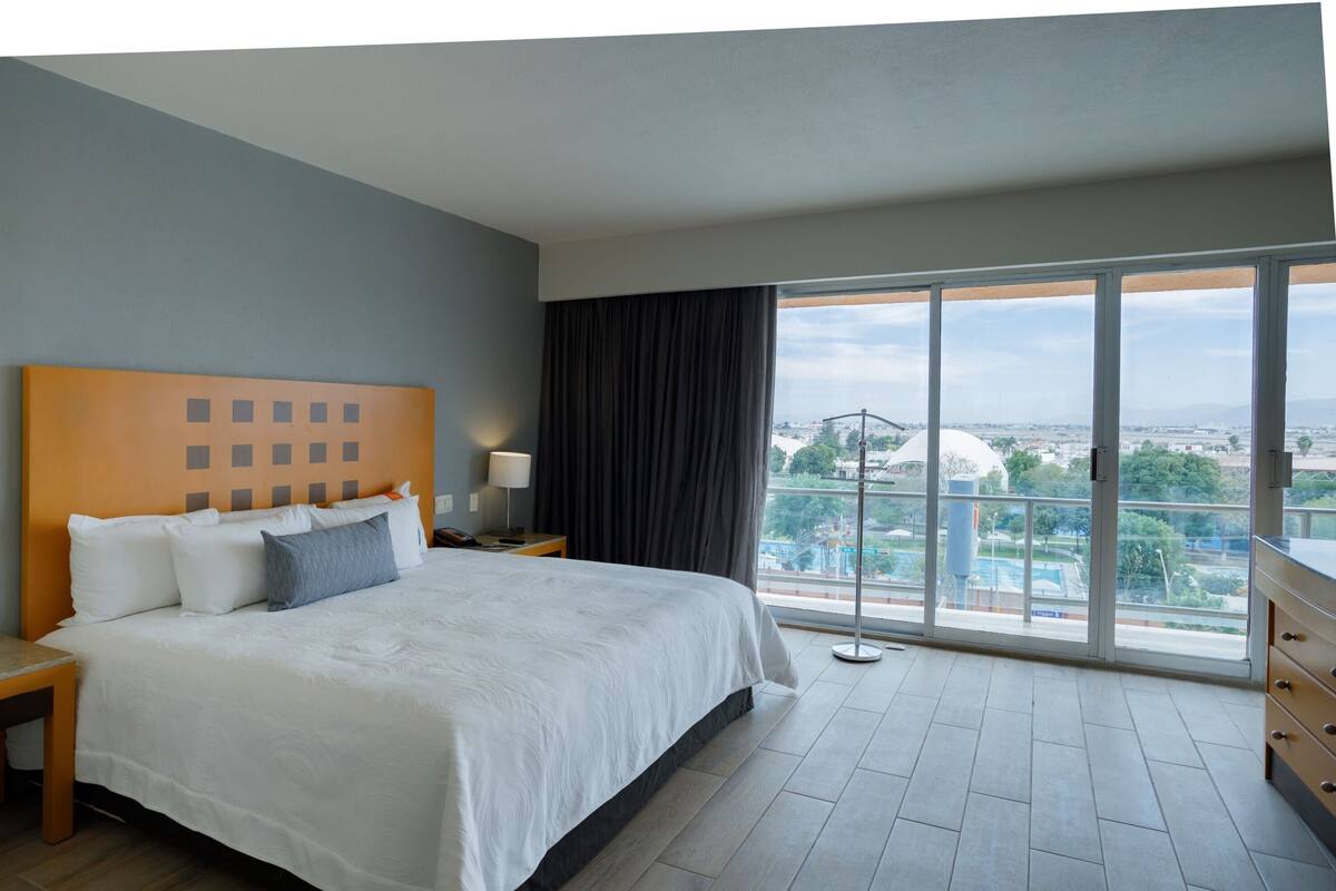 A hotel room with a large bed, a nightstand, a lamp, a view of a cityscape through large windows, and a balcony with a railing.