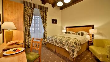 A hotel room with a bed, two bedside lamps, a wooden chair, a small table with a plate of fruit, and a window with curtains.