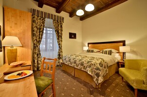 Een hotelkamer met een bed, twee bedlampen, een houten stoel, een klein tafeltje met een schaal fruit, en een raam met gordijnen.