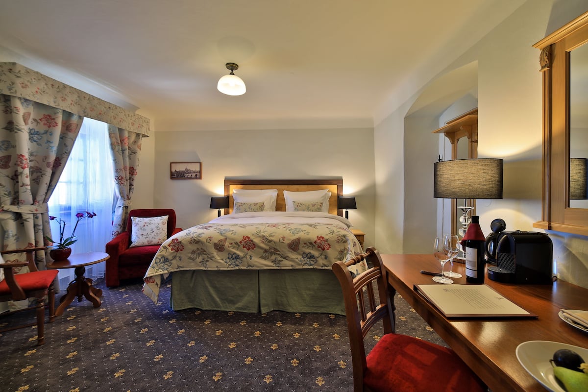 Habitación de hotel con cama, escritorio, silla, mesa, lámpara, una botella de vino y una planta.