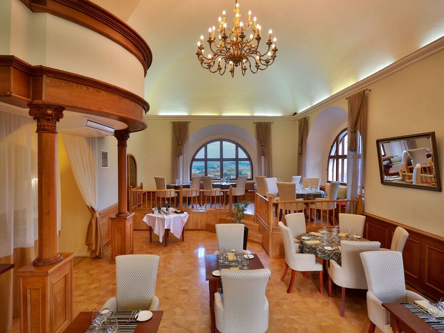 Un comedor de hotel con varias mesas y sillas, una araña y ventanas grandes con cortinas.