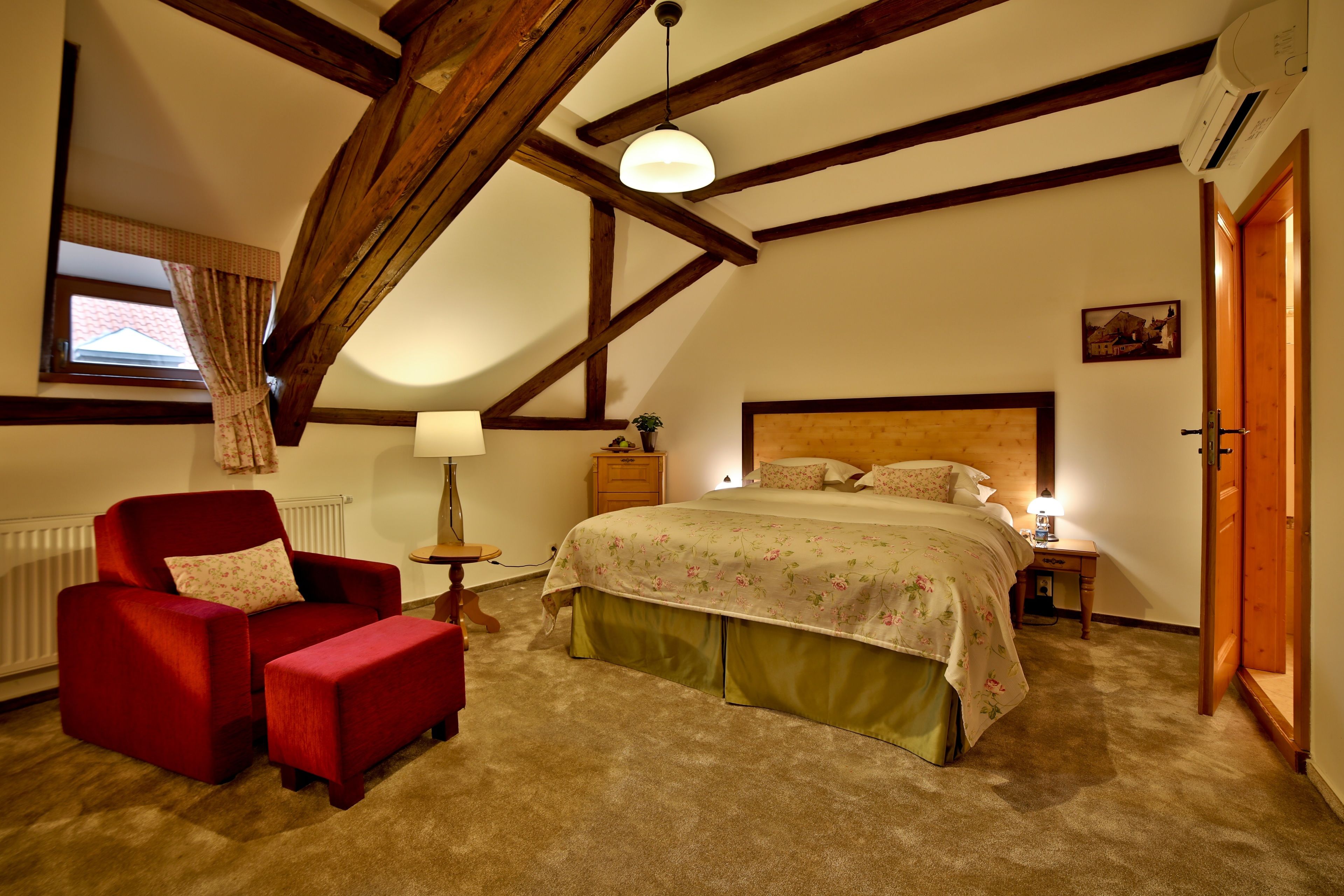 Ein Schlafzimmer mit schrägem Dach, einem Holzkopfteil, einem großen Bett mit floralem Bettbezug, einem roten Sessel und einem kleinen Nachttischchen mit Lampe.