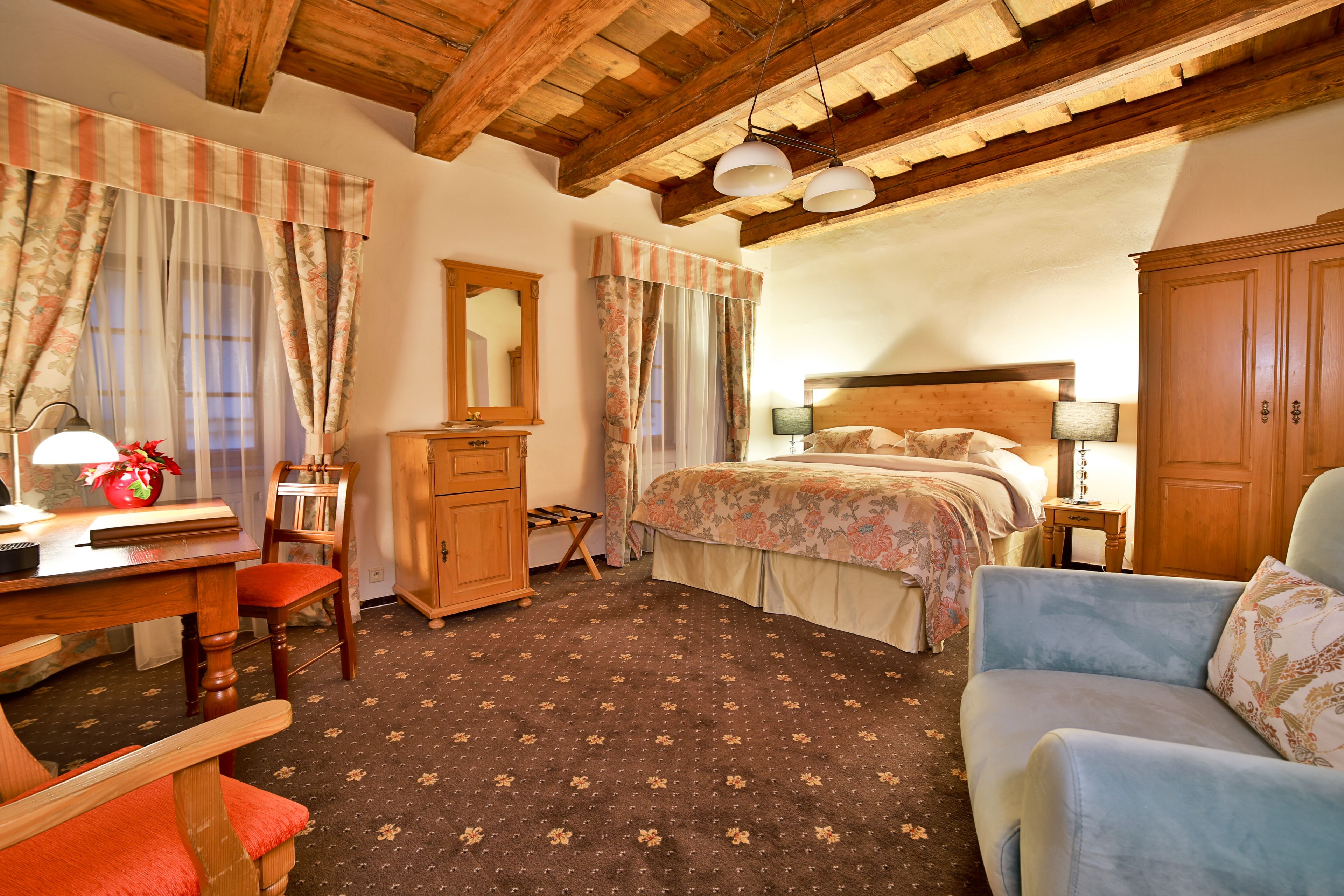 Une chambre avec un plafond en bois, un lit avec une literie à motifs floraux, un bureau avec une lampe, une chaise et une armoire.