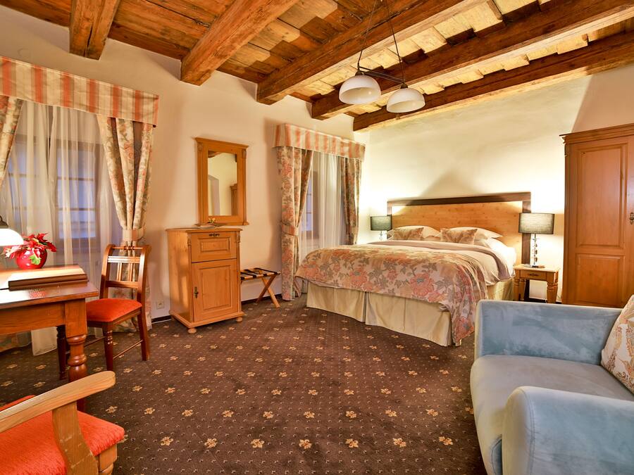 Une chambre avec un plafond en bois, un lit avec une literie à motifs floraux, un bureau avec une lampe, une chaise et une armoire.
