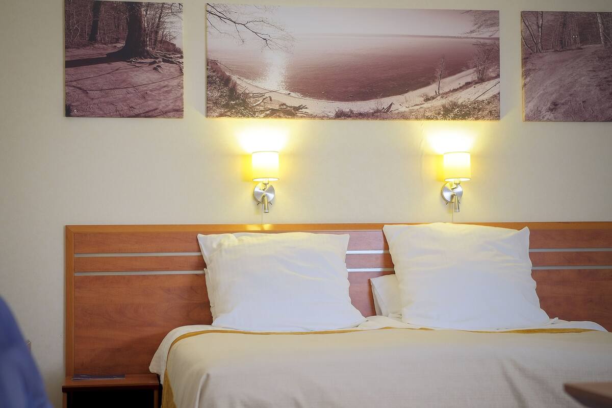 Habitación de hotel con cabecera de madera, dos almohadas blancas, una mesita de noche y dos lámparas de pared.