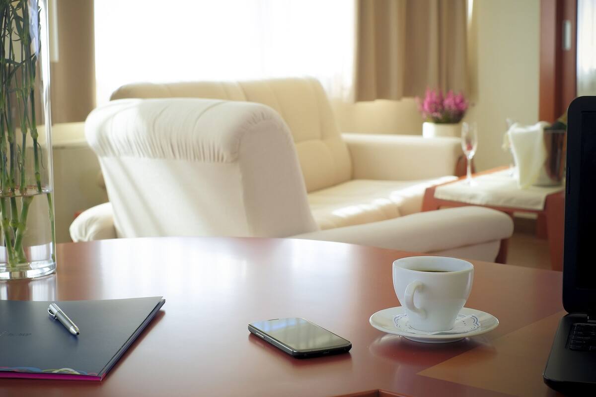 Una habitación con un sofá blanco, una mesa con una computadora portátil, una taza de café, un teléfono inteligente y un florero con flores.