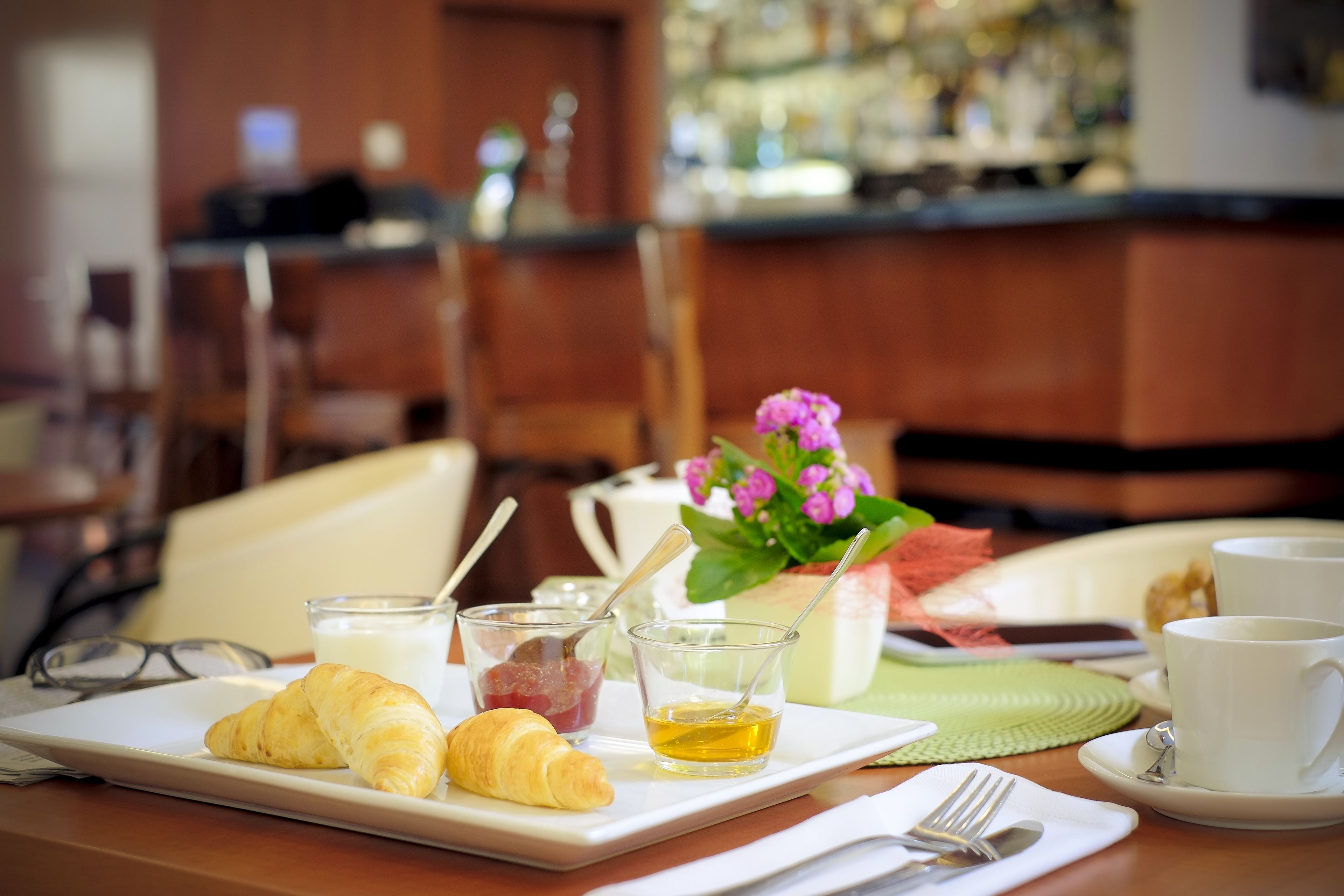 Disposição para o café da manhã com croissants, um copo de leite, uma xícara de chá e um arranjo de flores sobre uma mesa.