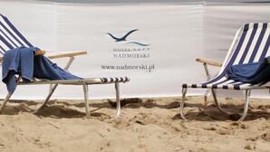 Duas cadeiras de praia com toalhas, um banner com logotipo e texto, e uma praia arenosa.