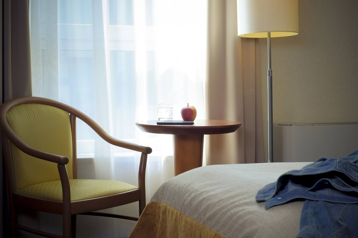 Habitación de hotel con una cama, una silla, una mesita con una manzana y un vaso, y una lámpara.