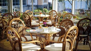 Um restaurante com cadeiras de vime e mesas redondas, grandes janelas com vista para a rua e carros estacionados.
