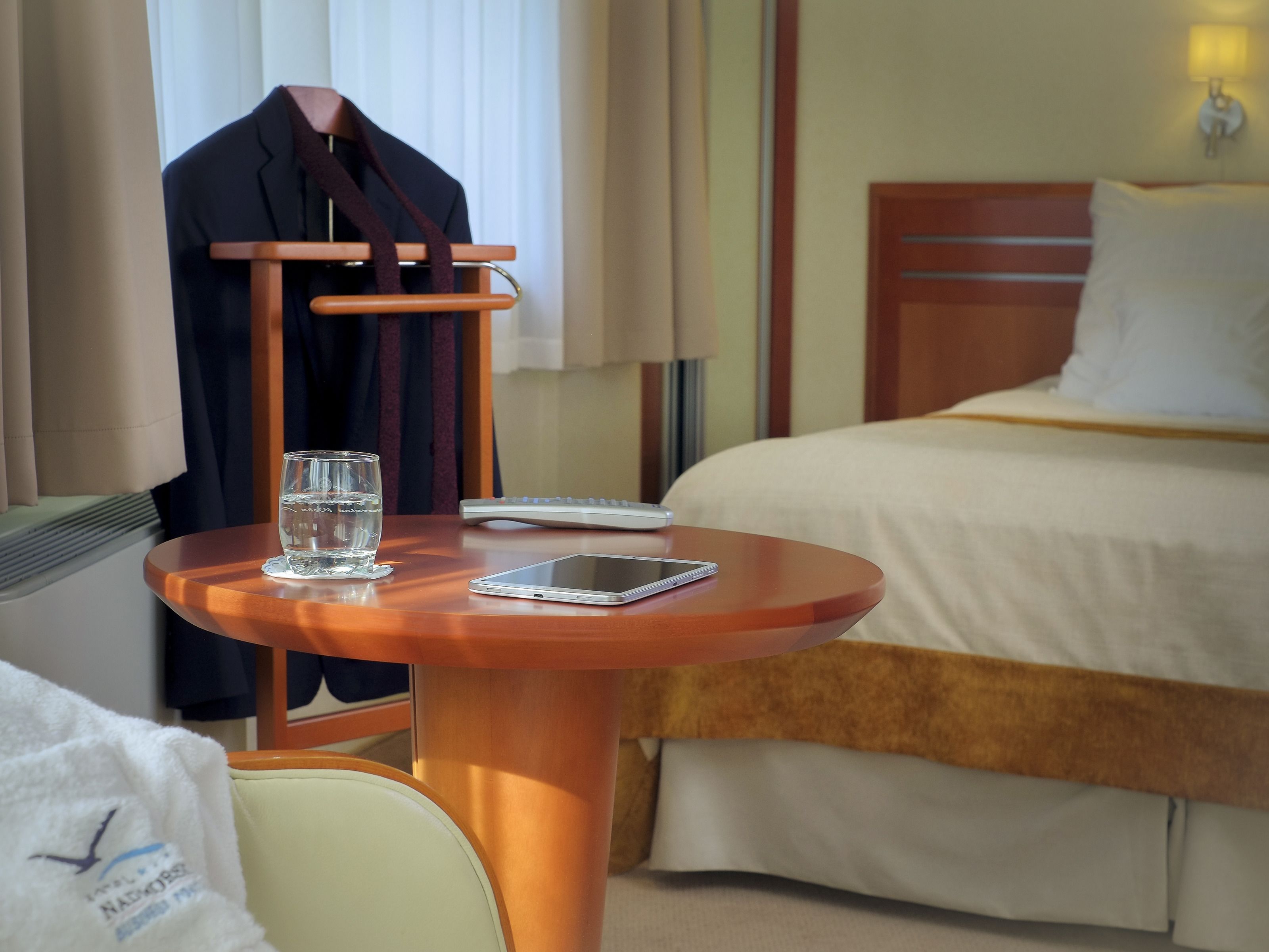 A hotel room with a bed, a bedside table with a glass of water and a smartphone, a hanging coat rack, and a lamp.