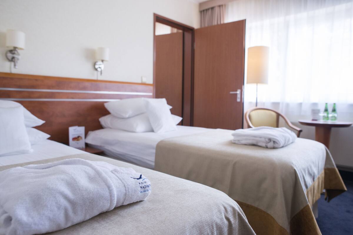 Habitación de hotel con dos camas, cabecero de madera, mesita de noche con lámpara y ventana con cortinas.