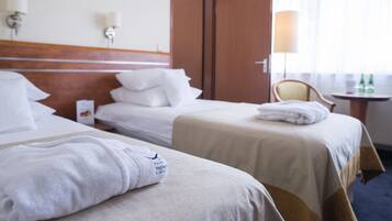 Habitación de hotel con dos camas, cabecero de madera, mesita de noche con lámpara y ventana con cortinas.