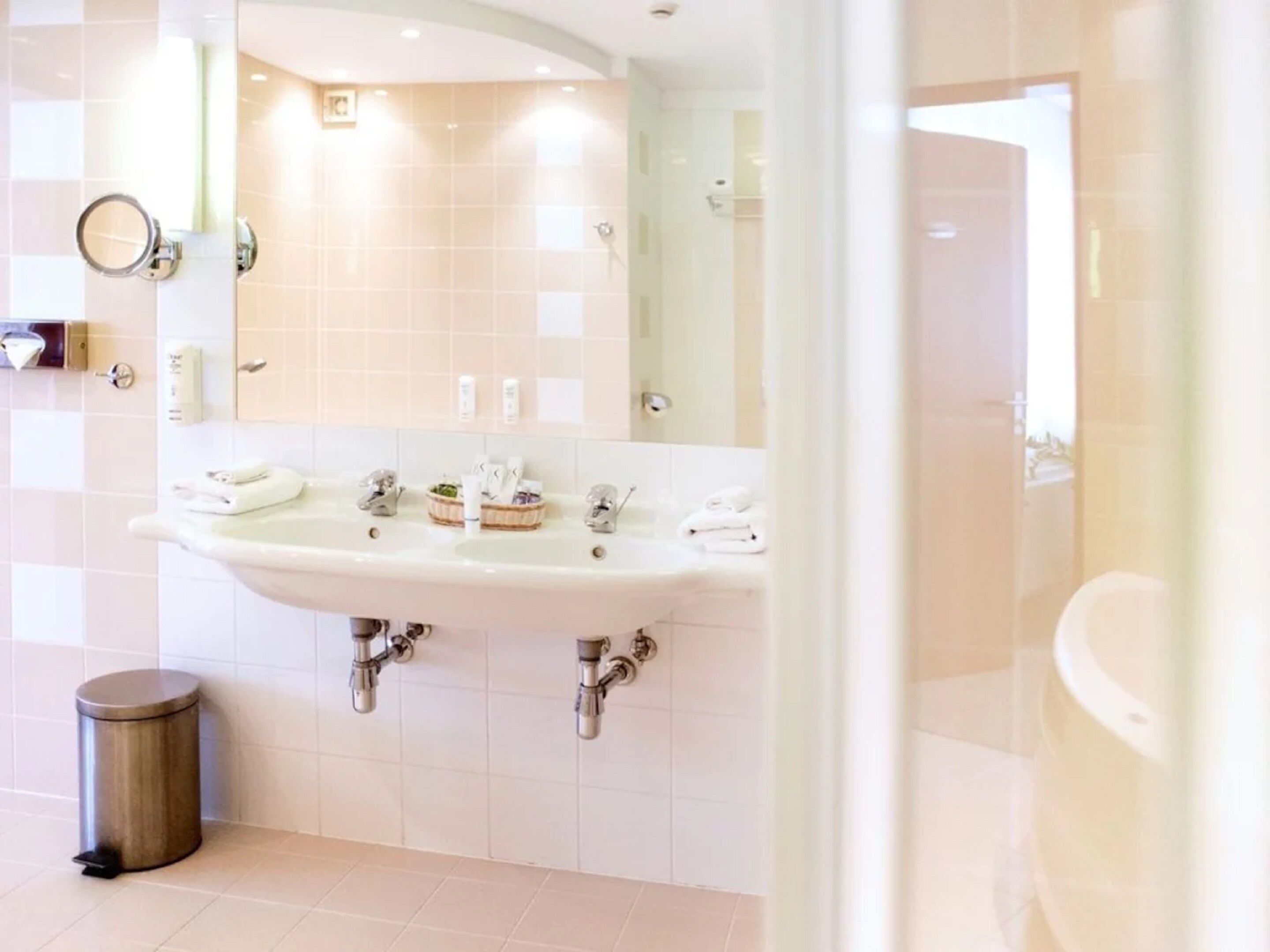 Une salle de bain moderne avec un grand lavabo, deux robinets et un miroir. On y trouve une poubelle et un porte-serviettes avec des serviettes.