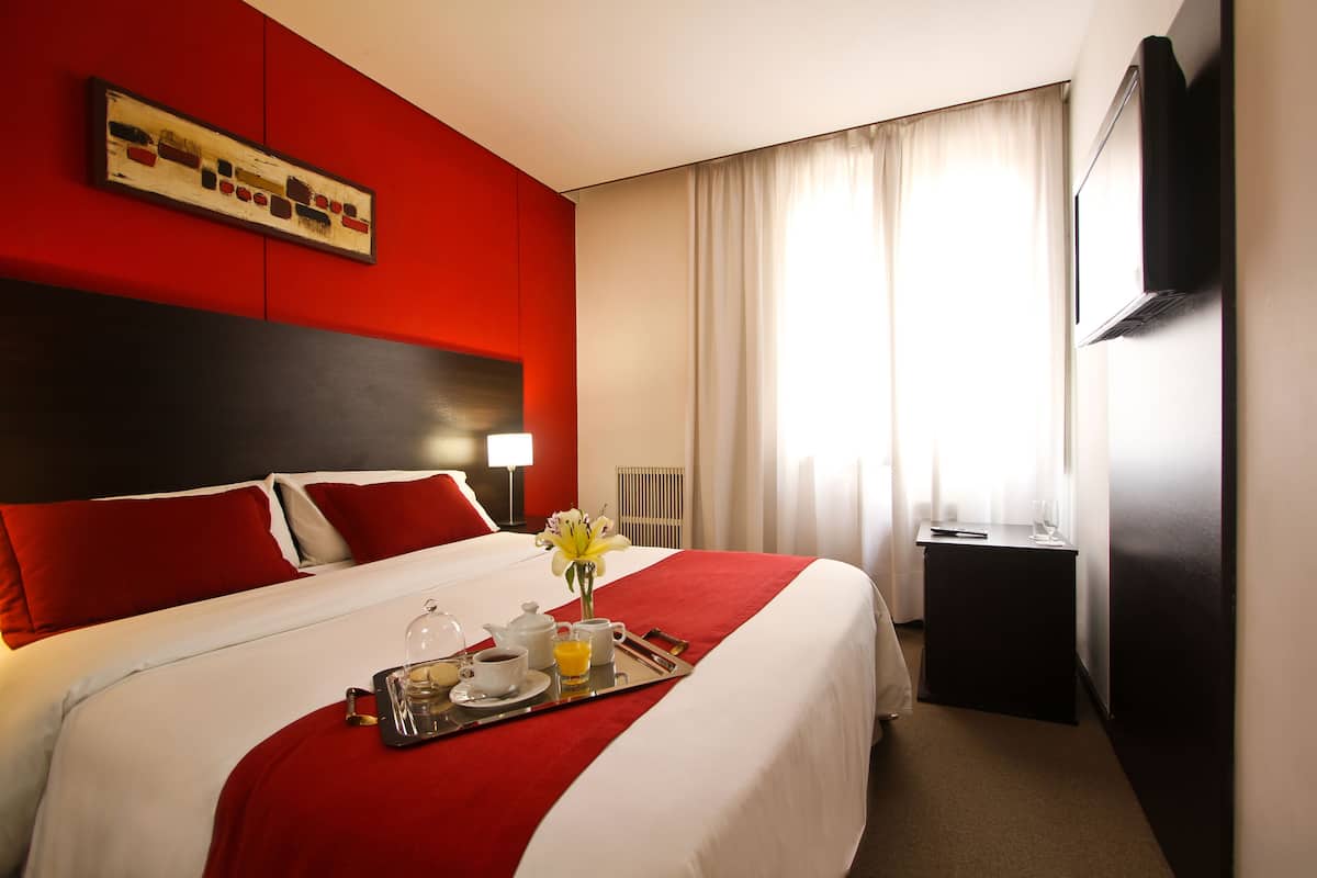 A hotel room with a large bed, red and white bedding, a tray with breakfast items, a vase with flowers, and a TV on the wall.