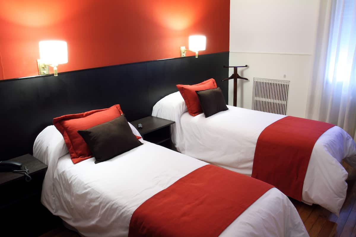 A hotel room with two beds, each with white bedding and red accents, a nightstand with a phone, and a wall-mounted light fixture.