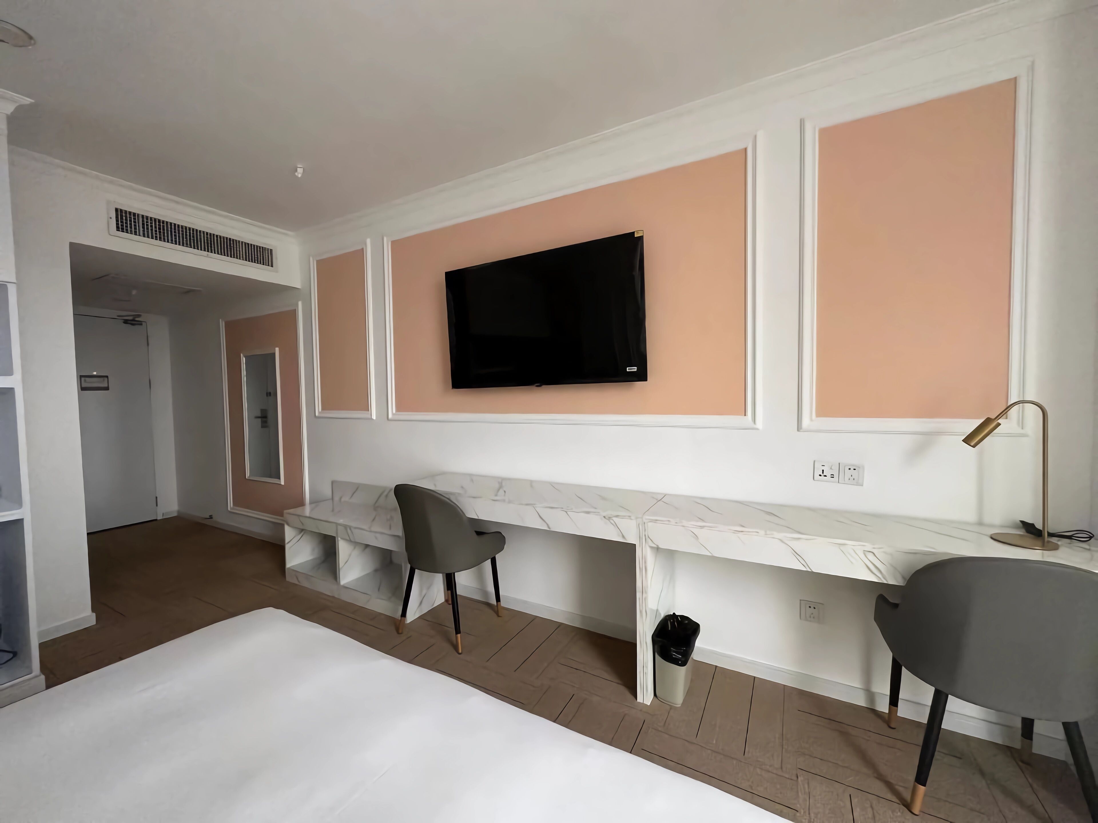 Une chambre d’hôtel moderne avec un bureau blanc, un téléviseur fixé au mur, une chaise grise et une petite poubelle.