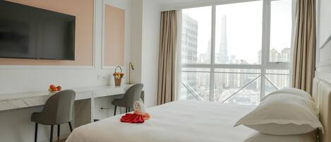 A hotel room with a bed, a desk, a chair, a flat-screen TV, and a city view through large windows.